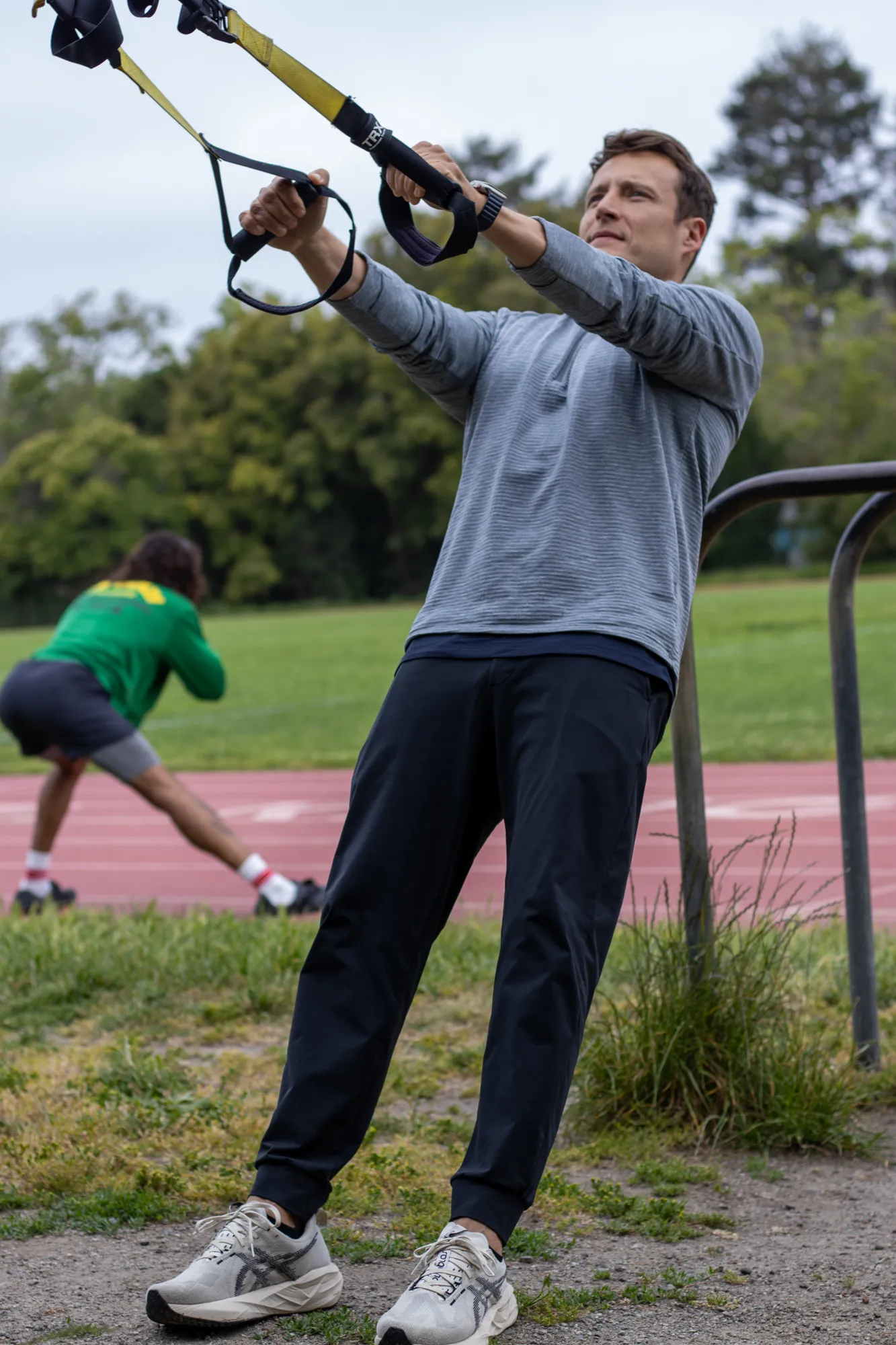 Dario coaching suspension-trainer work outdoors while a client stretches nearby